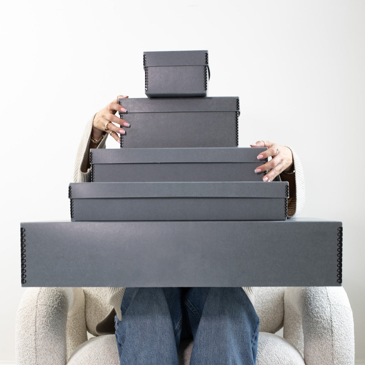 Someone sitting in a cream bouclé armchair holding a stack of grey Kept acid-free storage boxes