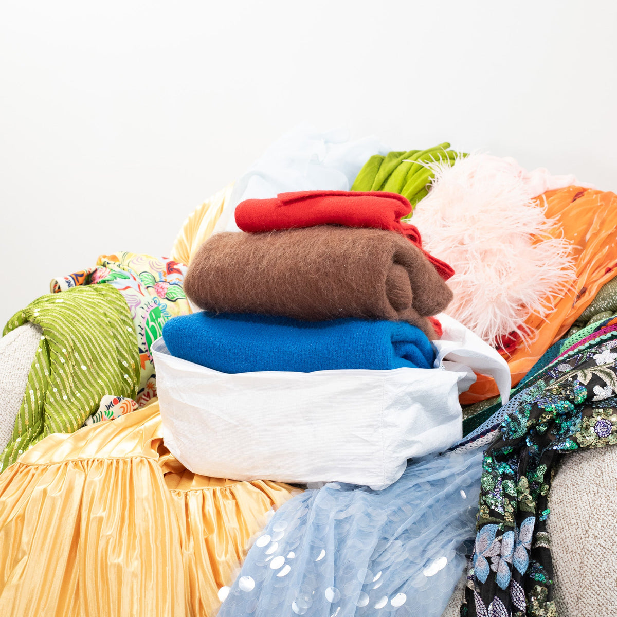 Folded rust, cream and blue jumpers stacked on a pile of colourful clothes on a bouclé armchair